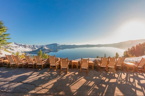 A row of wooden chairs overlooks a serene lake surrounded by snow-capped mountains under a clear blue sky.
