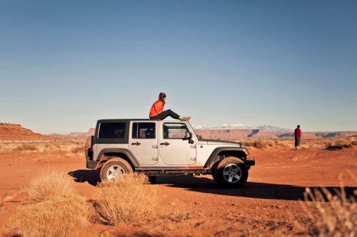 A person sits on the roof of a Jeep in a desert landscape, with another figure in the background and mountains in the distance.