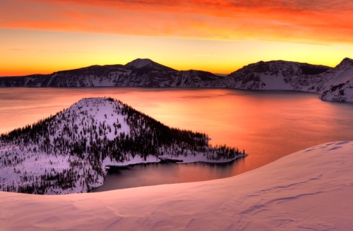 Sunset over a snowy landscape with a lake, mountains, and a small island, creating a serene winter scene.