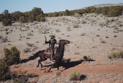 A cowboy on a rearing horse holds a pistol in a desert landscape with sparse vegetation.