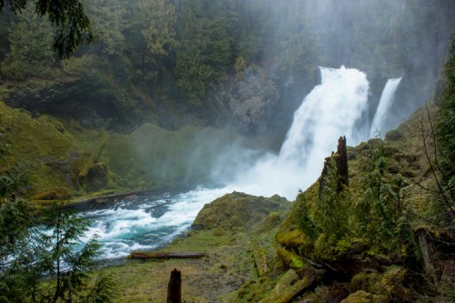 A misty waterfall cascades into a lush, green landscape surrounded by trees and moss-covered rocks.