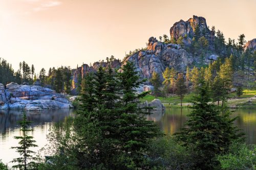 A serene landscape featuring a calm lake, rocky formations, and lush green trees under a warm sunset sky.