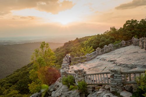 A scenic view from a rocky overlook at sunset, with lush green hills and a wooden railing in the foreground.