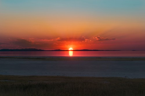 A vibrant sunset over a calm body of water, with colorful clouds and distant mountains silhouetted against the sky.