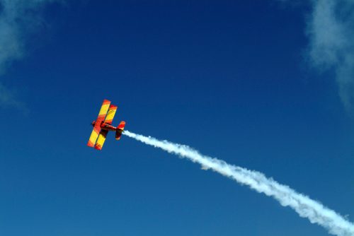 A colorful biplane performs a stunt, trailing white smoke against a clear blue sky.