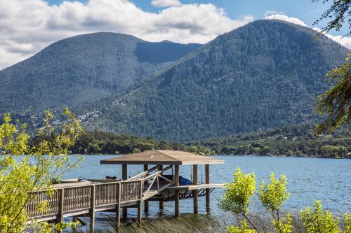 A serene lake view with a wooden dock, surrounded by lush green mountains under a partly cloudy sky.