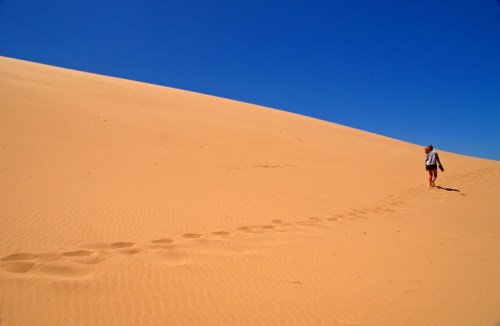 A person walks alone on a sandy dune under a clear blue sky, leaving footprints behind.