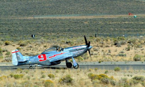 A vintage silver airplane with a red number 9 takes off from a desert runway, surrounded by sparse vegetation.