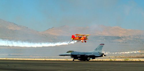 A red biplane performs a stunt above a gray F-16 jet on a runway, with smoke trails in the background and mountains nearby.