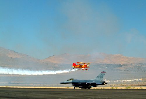 A red biplane performs a stunt above a parked F-16 fighter jet on an airfield, with mountains in the background.
