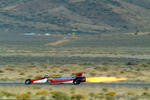 A jet-powered car speeds down a desert track, leaving a trail of flames behind it. Mountains are visible in the background.