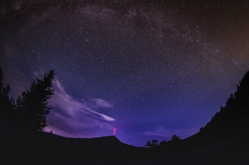 A panoramic night sky filled with stars, featuring a silhouette of trees and a hill with red-lit towers in the distance.