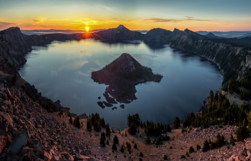 A panoramic view of Crater Lake at sunset, with a calm water surface and a small island surrounded by mountains.