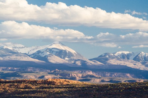 Snow-capped mountains under a blue sky with fluffy clouds, showcasing a rugged landscape and distant peaks.