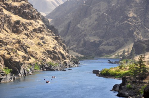 A serene river flows through rocky, dry hills, with a few kayakers paddling along the calm water.