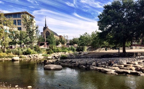A serene park scene with a pond, rocks, trees, and a church in the background under a blue sky with clouds.