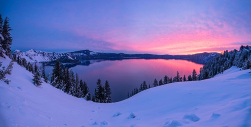 A serene winter landscape at Crater Lake, with snow-covered mountains and a vibrant pink and purple sunset reflecting on the water.