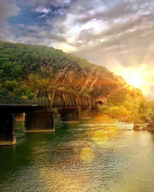 A scenic view of a bridge over a river, surrounded by lush hills and a vibrant sunset in the background.