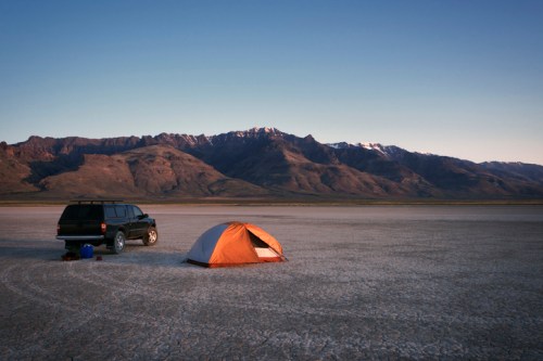 A tent and a parked SUV on a dry, cracked salt flat with mountains in the background during sunset.
