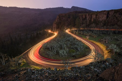 A winding road curves through a mountainous landscape, illuminated by car lights at dusk.