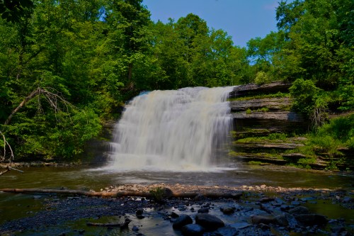 A serene waterfall cascades over rocky ledges, surrounded by lush green trees and a calm river below.