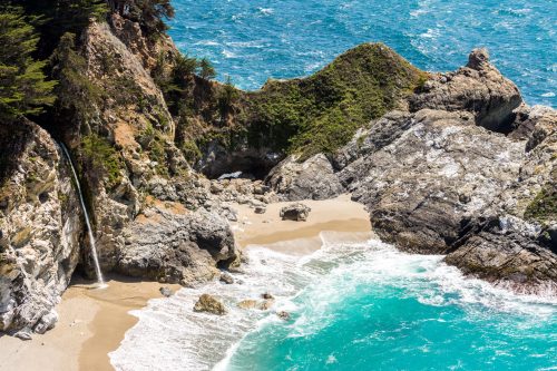 A scenic view of a waterfall cascading onto a sandy beach, surrounded by rocky cliffs and vibrant blue ocean waves.