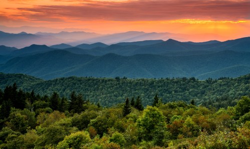 A scenic view of rolling mountains at sunset, with layers of green trees and a colorful sky.