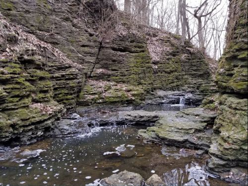 A serene rocky landscape with moss-covered cliffs and a small stream flowing into a calm pool of water.