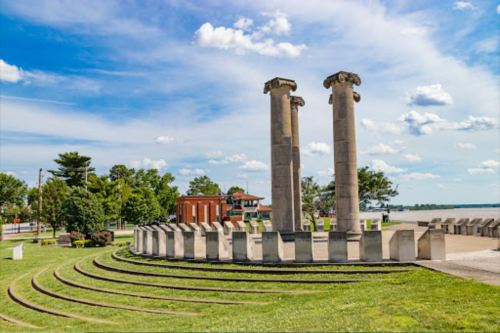 Two tall stone columns stand amidst a grassy area with a circular stone seating arrangement and a blue sky in the background.