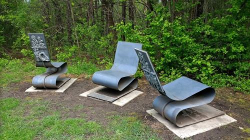 Three modern metal sculptures resembling lounge chairs, set in a wooded area with greenery in the background.