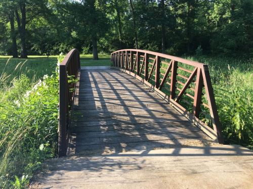A wooden bridge with a metal railing spans a grassy area, casting shadows on the path below. Lush greenery surrounds it.