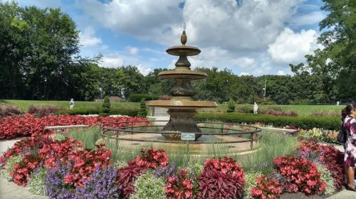 A tiered fountain surrounded by colorful flowers and greenery under a partly cloudy sky.