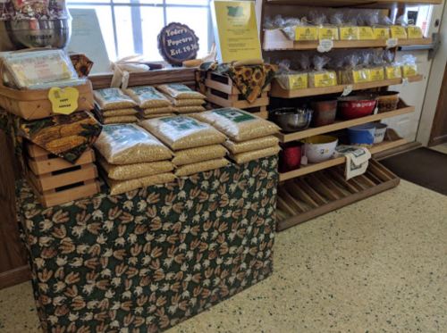 A display of bags of popcorn kernels on a table, with shelves of snacks and containers in the background.