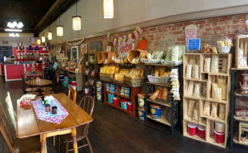 A cozy shop interior with wooden shelves filled with snacks, popcorn, and a dining table in the foreground.
