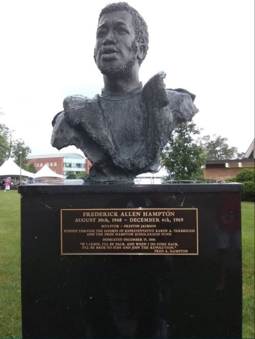 A bust of Frederick Allen Hampton with an inscription detailing his life and dedication date, set in a park.