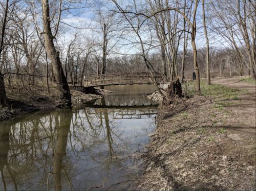A serene landscape featuring a calm creek, a wooden bridge, and bare trees under a blue sky.