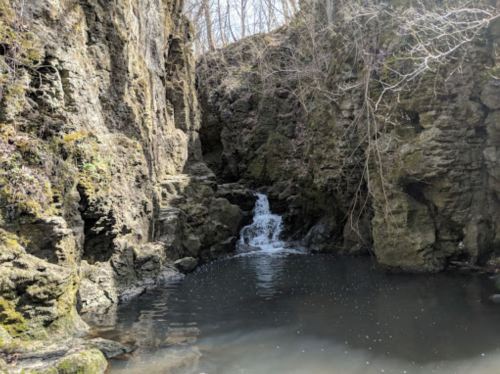 A serene rocky landscape with a small waterfall cascading into a calm pool surrounded by trees.