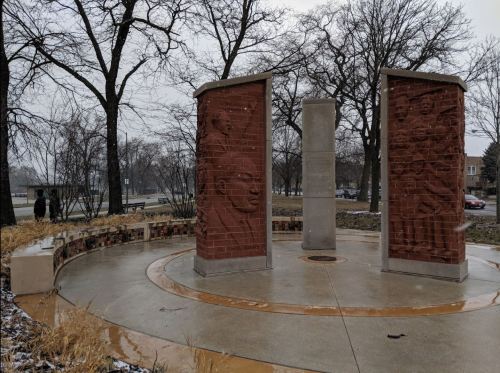 A circular memorial with two brick pillars featuring relief sculptures, surrounded by trees in a snowy park.