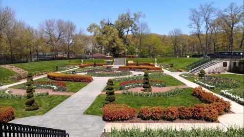 A beautifully landscaped garden with colorful flower beds, pathways, and trees under a clear blue sky.