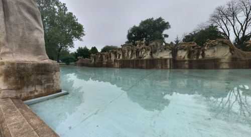 A serene, reflective pool with a sculpted stone wall and trees in the background, under a cloudy sky.