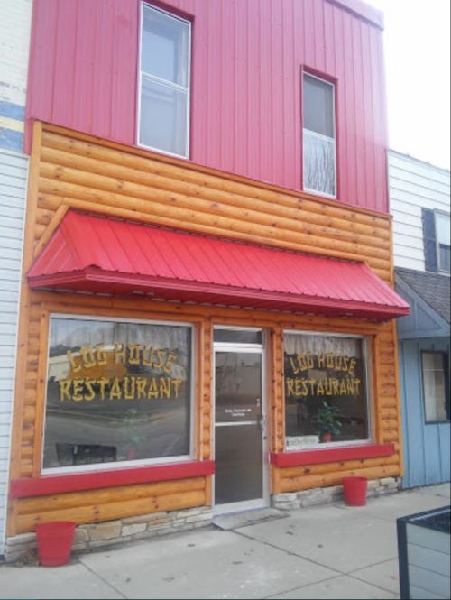 Exterior of a log cabin-style restaurant with a red metal roof and large windows displaying "Log House Restaurant."