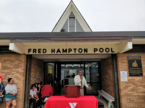 A speaker addresses a crowd at the entrance of Fred Hampton Pool, with a red tablecloth in front.