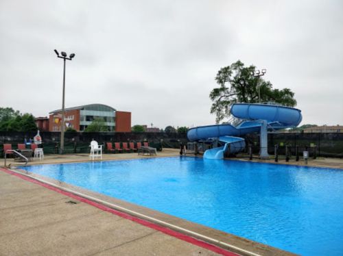 A clear blue swimming pool with a water slide, surrounded by lounge chairs and a cloudy sky.