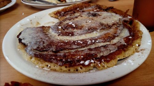 A plate of cinnamon roll pancakes topped with powdered sugar and syrup, served in a casual dining setting.