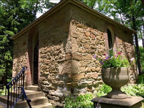 A small stone building with a sloped roof, surrounded by greenery and a flower pot on the steps.