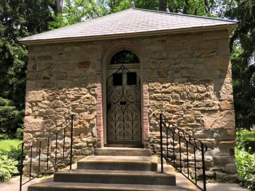 A small stone building with a sloped roof, featuring a decorative door and steps leading up to it, surrounded by greenery.