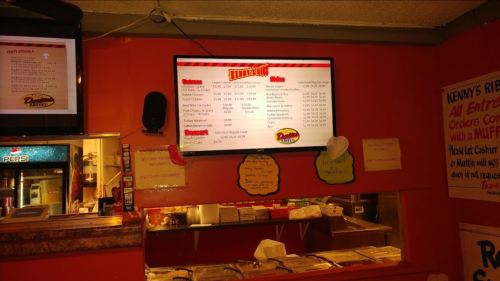 Menu displayed on a screen in a restaurant, featuring various food items and prices. Brightly colored walls in the background.