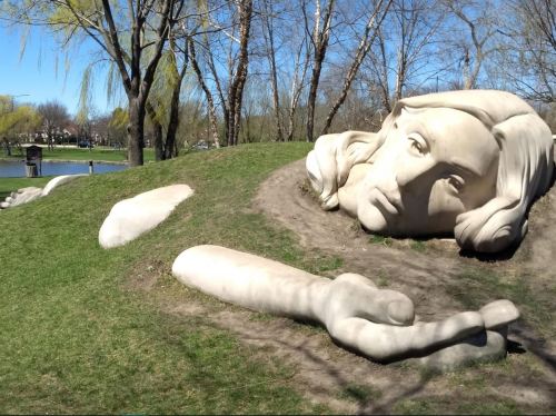 A large, sculpted face and arm emerging from a grassy hill near a pond, surrounded by trees on a sunny day.