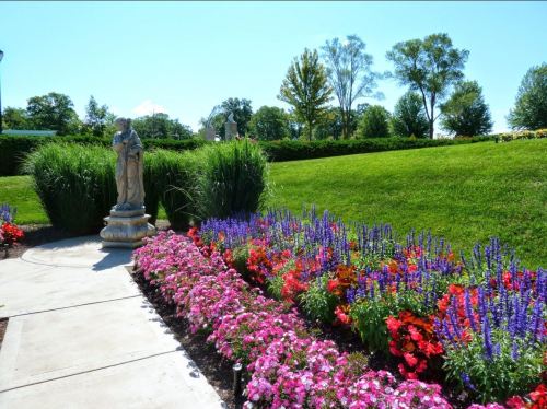 A garden path lined with colorful flowers and a statue, surrounded by lush greenery under a clear blue sky.