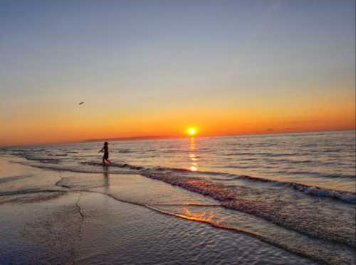 A person walks along the shoreline at sunset, with vibrant orange and yellow hues reflecting on the water.